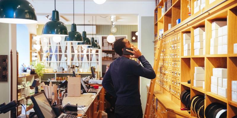 Man on a phone stands at a wooden-shelved retail shop counter stocked with boxed products