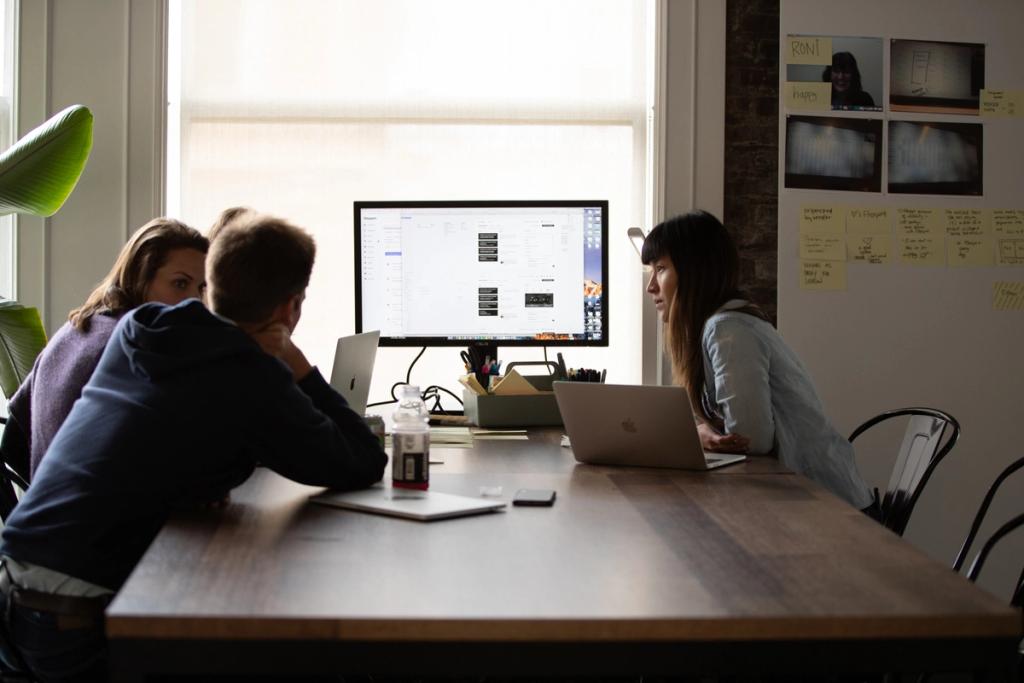 Three colleagues at a wood table discussing software on a large monitor in a sunlit office