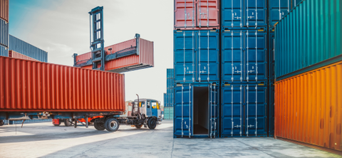 Photo of a container yard with a reach stacker lifting a red container above a truck chassis, beside stacks of blue and orange containers.