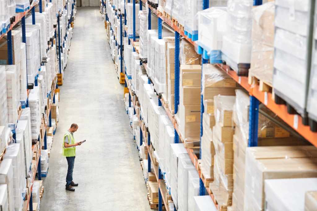 Worker in a hi-vis vest checking a tablet in a long aisle of a stocked warehouse