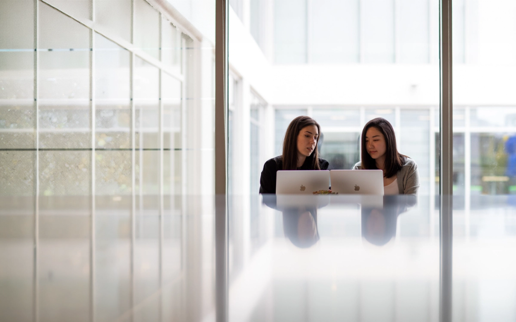 Two women collaborating on a laptop while seated at a table in a modern office setting.