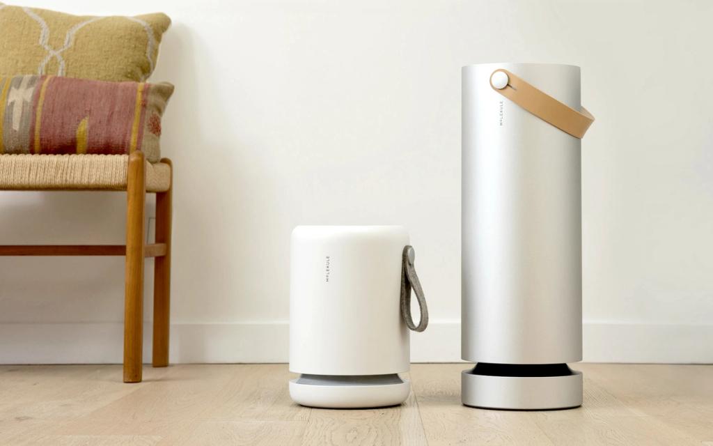 White and silver Molekule air purifiers standing side by side beside a chair with pillows