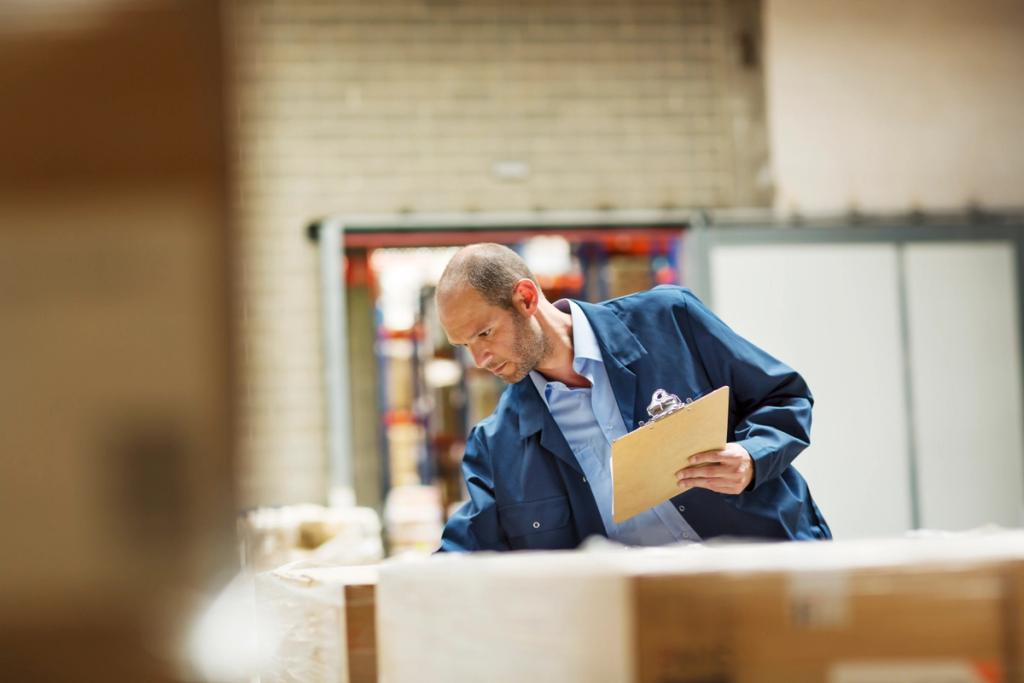 Warehouse worker in blue coveralls reviewing a clipboard beside palletized goods, with shelving and freight blurred behind