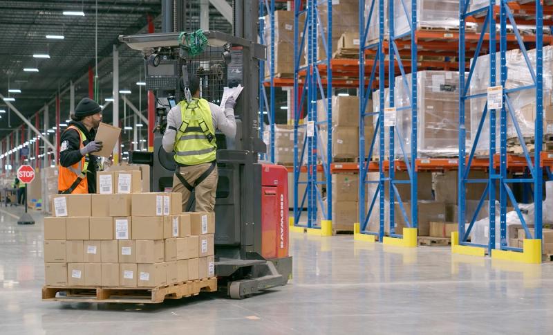 Dallas fulfillment center with workers loading a pallet onto a reach forklift beside tall racking