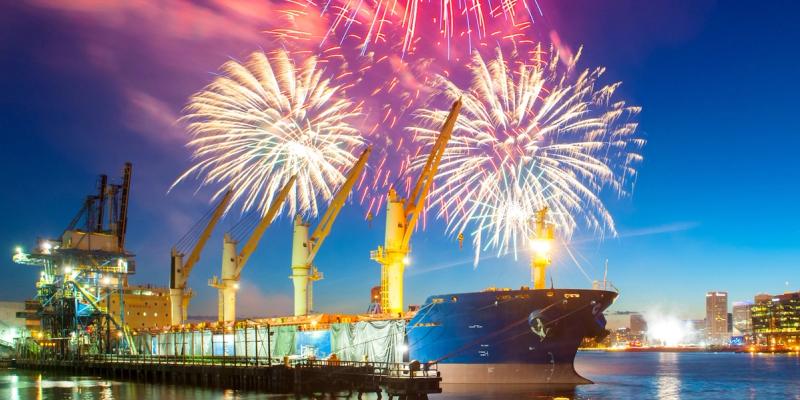 Fireworks burst over a cargo ship and harbor cranes at dusk with a city skyline in the background.