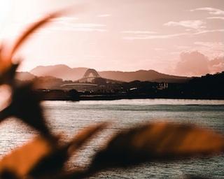 Photo across calm water toward an arched bridge and low hills at dusk, framed by a blurred leaf in the foreground.