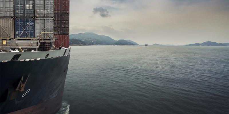 Bow of a container ship stacked with containers sailing on grey water toward distant hills