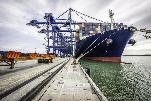 Container ship moored at a port quay beside blue gantry cranes, viewed along the dock