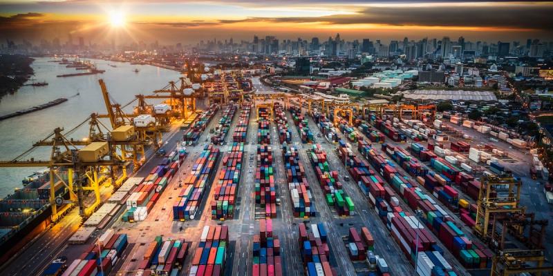Aerial view of a busy container port with cranes and stacked containers at sunset before a city skyline