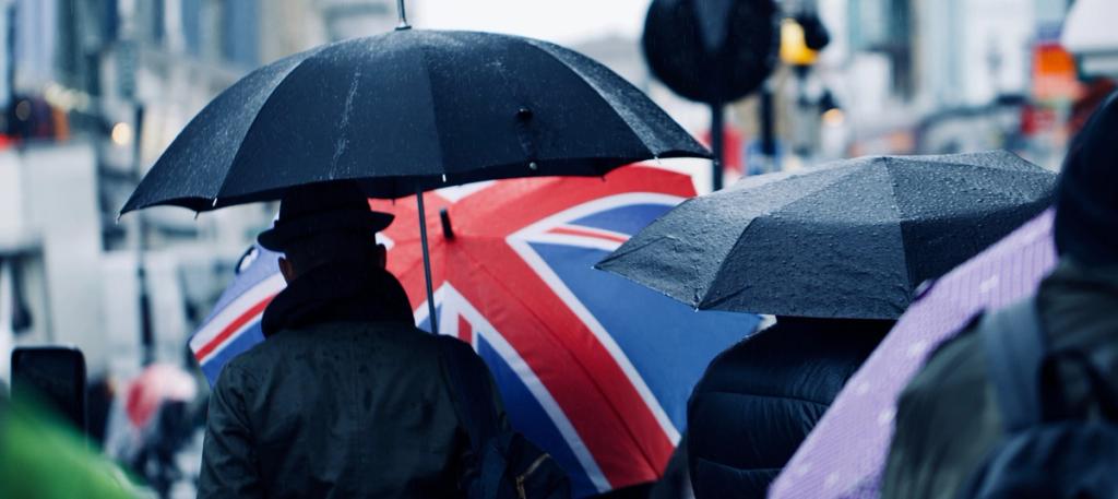 Pedestrians with black umbrellas and a Union Jack umbrella walk through rain on a city street