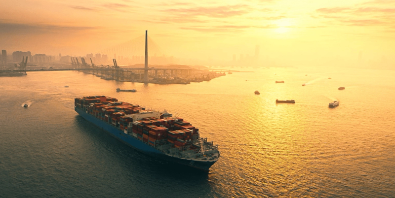 Container ship sailing past a city skyline and bridge at golden sunset, smaller vessels nearby