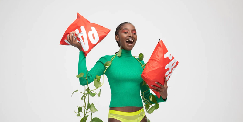 Smiling woman in a green top holding two red compostable mailer bags against a white backdrop