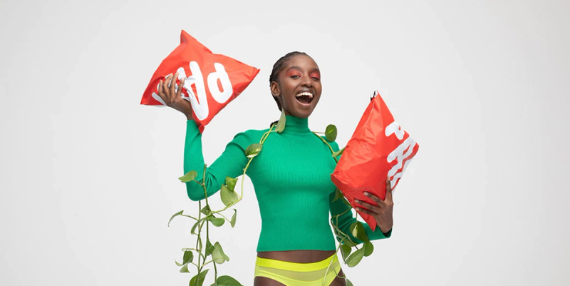 Smiling woman in a green top holding two red compostable mailer bags against a white backdrop