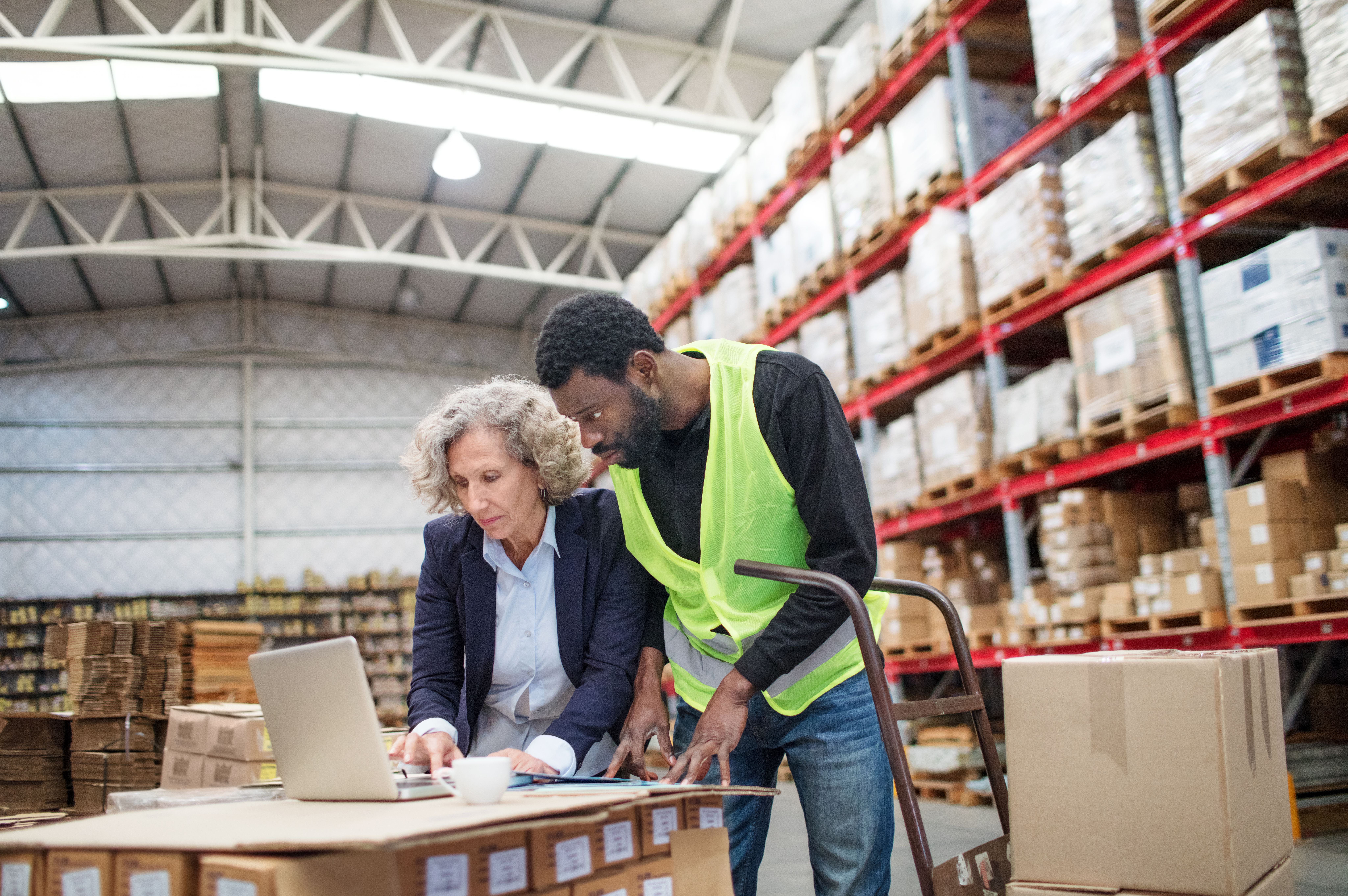 Warehouse worker in safety vest assists female manager reviewing documents at a table.
