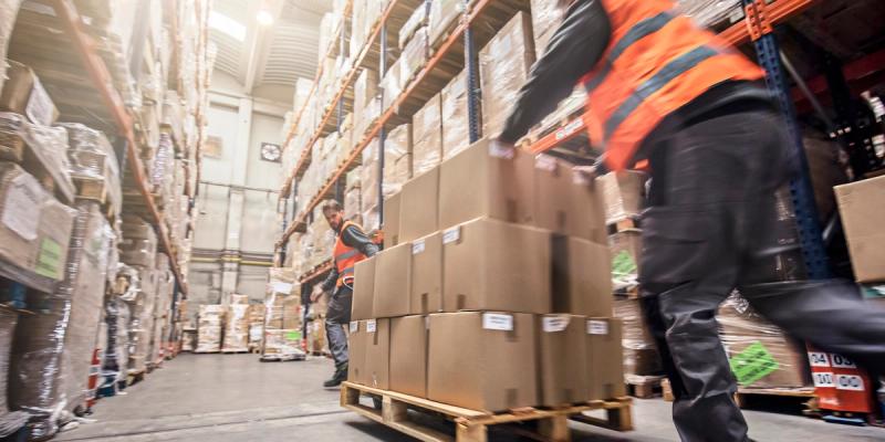 Workers in orange vests move pallets of cardboard boxes through a busy warehouse aisle, motion-blurred