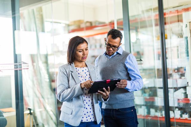 Woman and man in business attire reviewing a tablet together in front of warehouse shelving