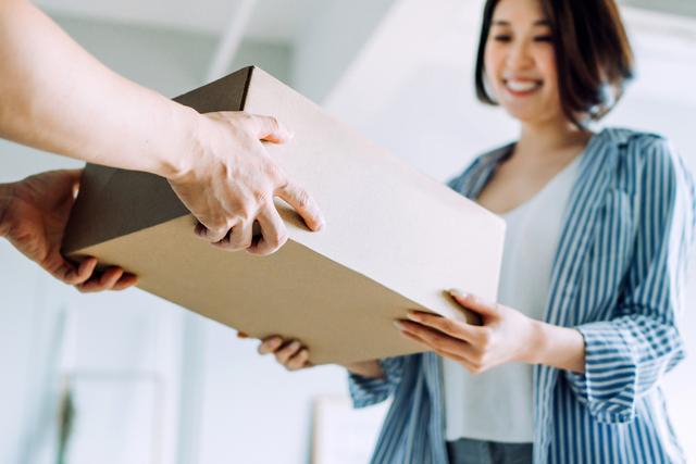 Hand passes a plain cardboard box to a smiling woman in a striped blue shirt indoors