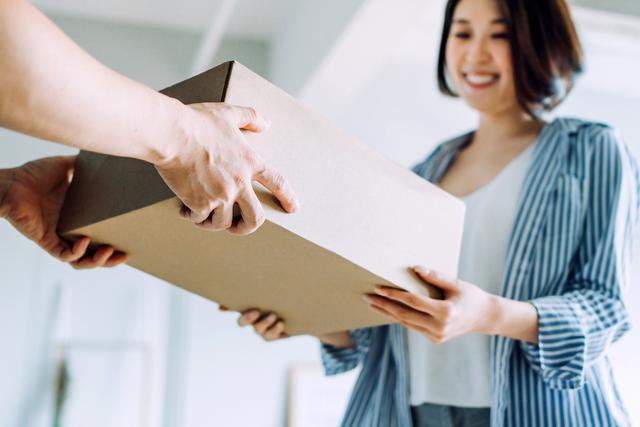 Hand passes a plain cardboard box to a smiling woman in a striped blue shirt indoors