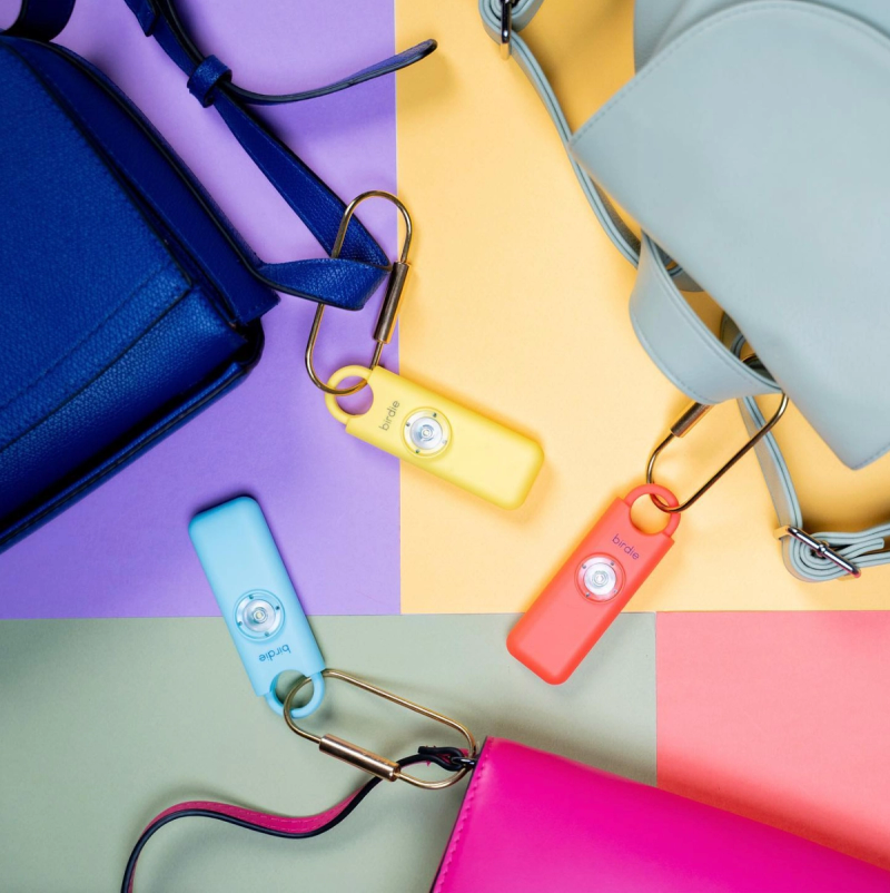 She's Birdie safety alarms in pastel colors clipped to handbags on a multicolored flat-lay