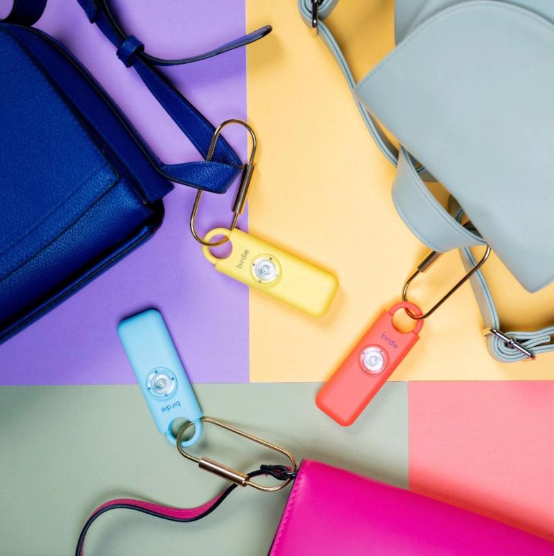 She's Birdie safety alarms in pastel colors clipped to handbags on a multicolored flat-lay