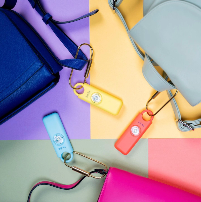 She's Birdie safety alarms in pastel colors clipped to handbags on a multicolored flat-lay