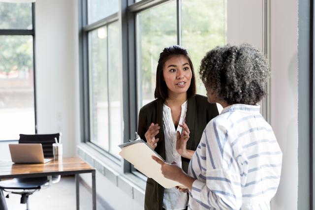 Two women talk by an office window, one holding a folder while the other gestures.