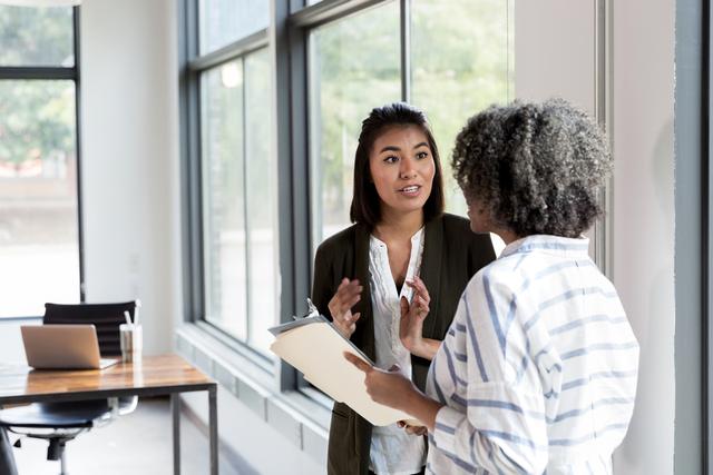 Two women talk by an office window, one holding a folder while the other gestures.