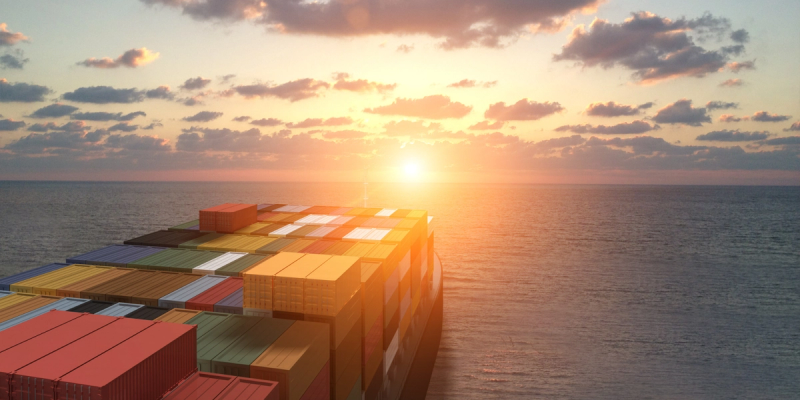 Rows of colorful shipping containers stacked on a vessel at sea with the sun low on the horizon