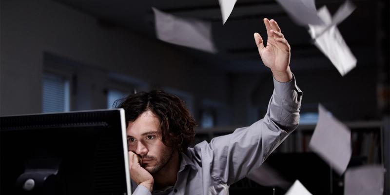 Frustrated man in a grey shirt throws papers into the air at his desk in a dark office
