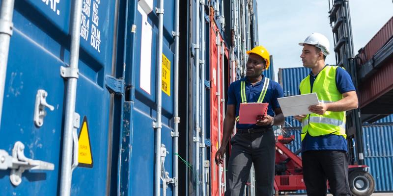 Two dock workers in hard hats and safety vests talk while walking between stacked shipping containers at a port.