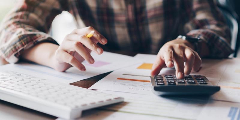 Close-up of a person in plaid shirt pressing a calculator over financial charts beside a keyboard