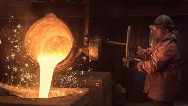 Foundry worker in a helmet and face shield pours glowing molten metal from a ladle, sparks flying in a dark workshop