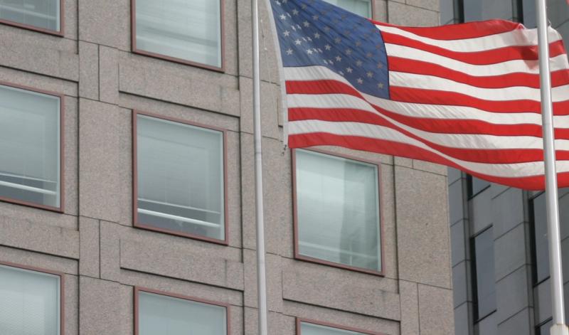 American flag waving on a pole in front of a gray stone office building with rows of windows
