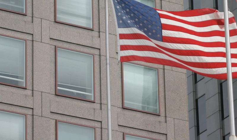 American flag waving on a pole in front of a gray stone office building with rows of windows