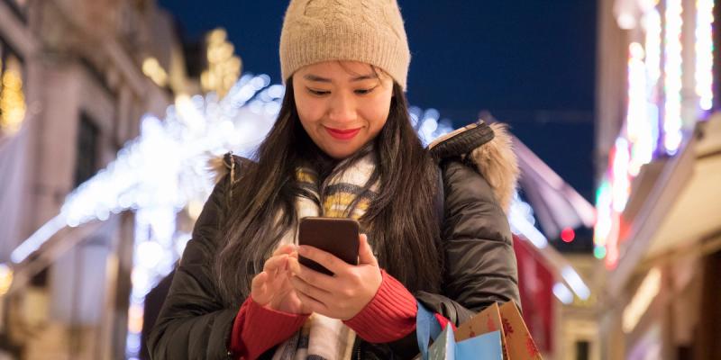 Smiling woman in knit hat and scarf reading her phone with shopping bags on a lit-up city street at night