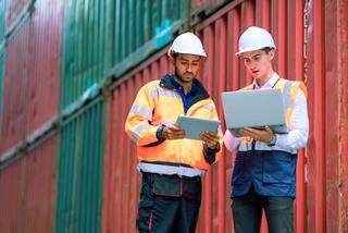 Two workers in hard hats and hi-vis vests reviewing a tablet and laptop in front of red shipping containers