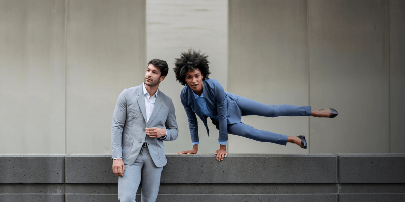 Man in gray suit leaning on a wall as woman in blue suit leaps horizontally beside him