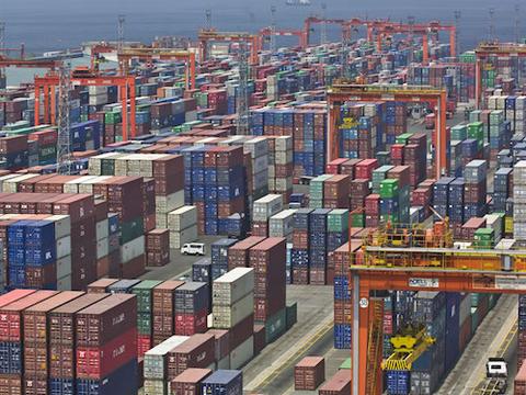 Aerial view of a container yard with thousands of stacked shipping containers in rows, served by orange gantry cranes alongside the water.