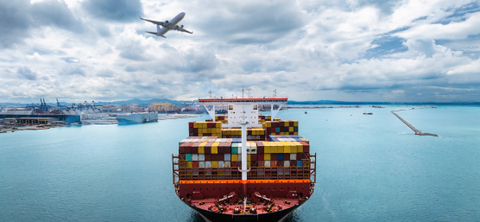 Aerial photo of a fully loaded container ship heading in toward a port terminal with a jet airliner flying overhead.