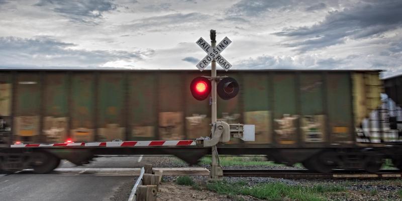 Freight train passing a railroad crossing with a flashing red signal and lowered striped gate under cloudy sky