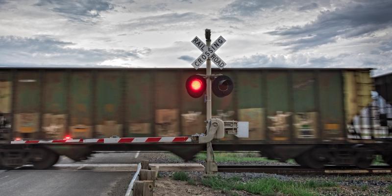 Freight train passing a railroad crossing with a flashing red signal and lowered striped gate under cloudy sky