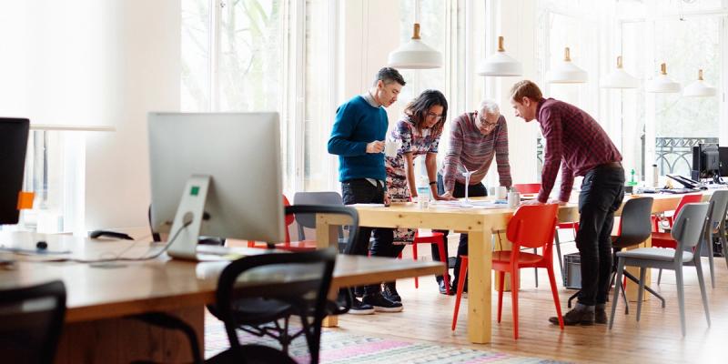 Four colleagues lean over a wooden table reviewing papers in a bright open-plan office