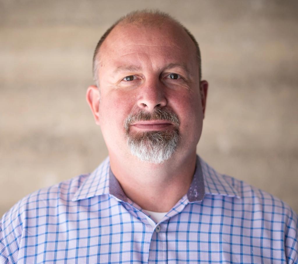 Headshot of a bald man with a gray goatee in a blue checked shirt against a neutral background.