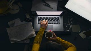 Overhead view of a person in a yellow sweater typing on a laptop at a dark desk while holding a cup of coffee