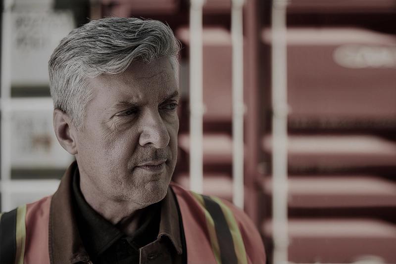 Gray-haired worker in a hi-vis vest looking aside in front of a stacked red shipping container