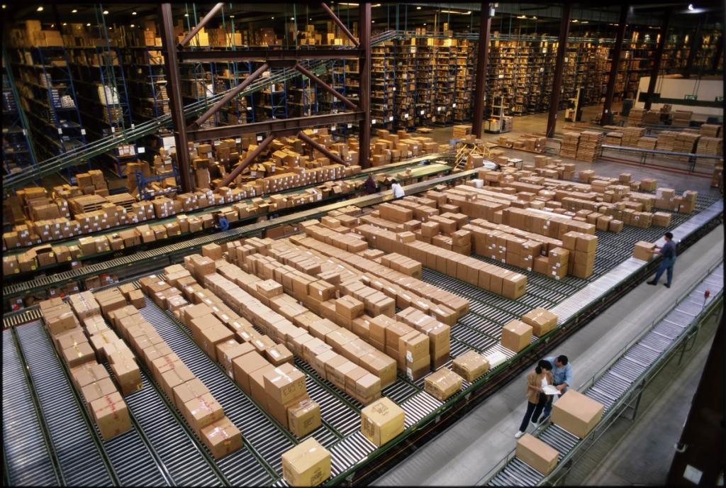 Warehouse aisles stacked with boxed inventory feeding rows of conveyor belts