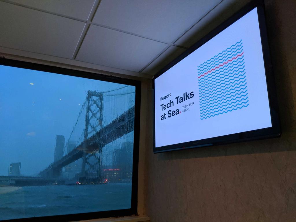 Wall-mounted screen displaying Flexport Tech Talks at Sea slide beside a window overlooking a suspension bridge