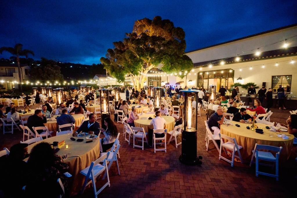 Outdoor evening reception with guests at round tables, string lights and tall patio heaters