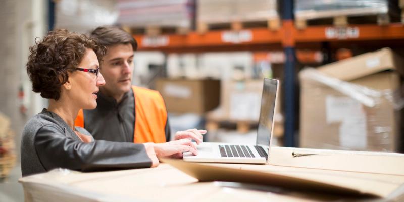 Businesswoman and a man in an orange hi-vis vest look at a laptop atop boxes in a warehouse