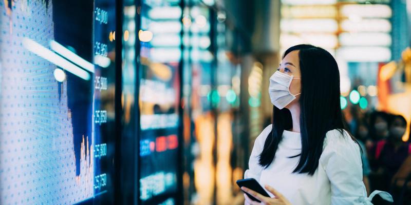 Woman in a face mask holding a phone, looking at a blue digital financial display board in a transit hall